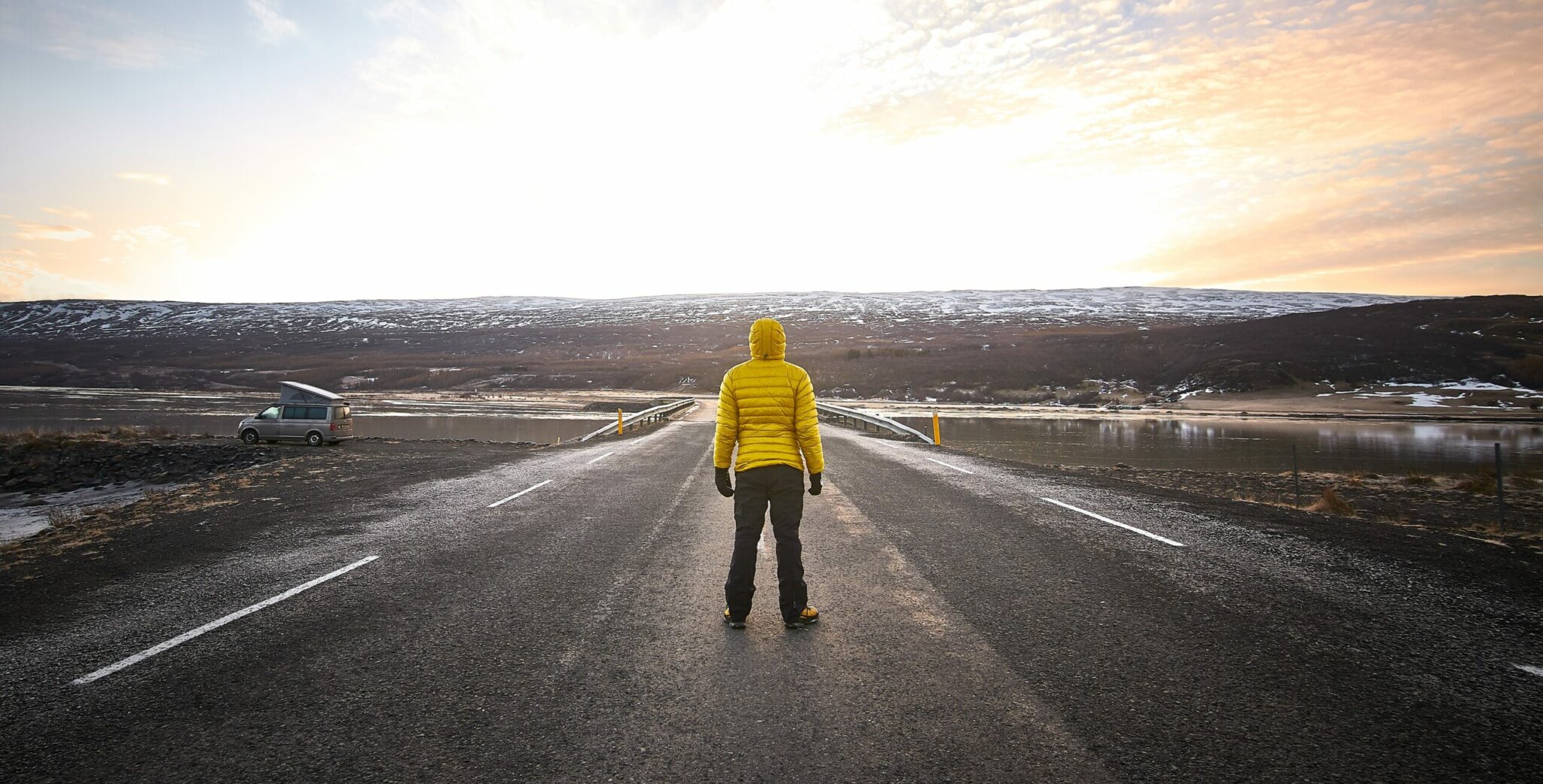 A male wearing a yellow jacket while standing in the middle of an empty road looking in the distance